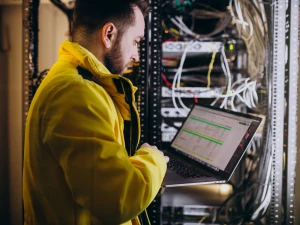 Platzhalterbild für die Rubrik it.service; es zeigt einen IT Techniker in gelber Jacke der vor einem Netzwerkschrank an einem Notebook arbeitet